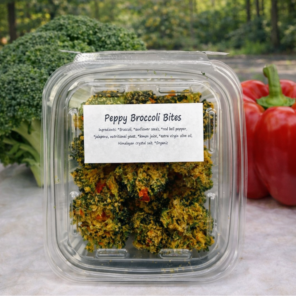 A clear plastic container of Healing Home Foods Peppy Broccoli Bites sits outdoors, with fresh broccoli and two red bell peppers behind it. The bites inside show broccoli and red pepper pieces, coated in sunflower seed sauce.