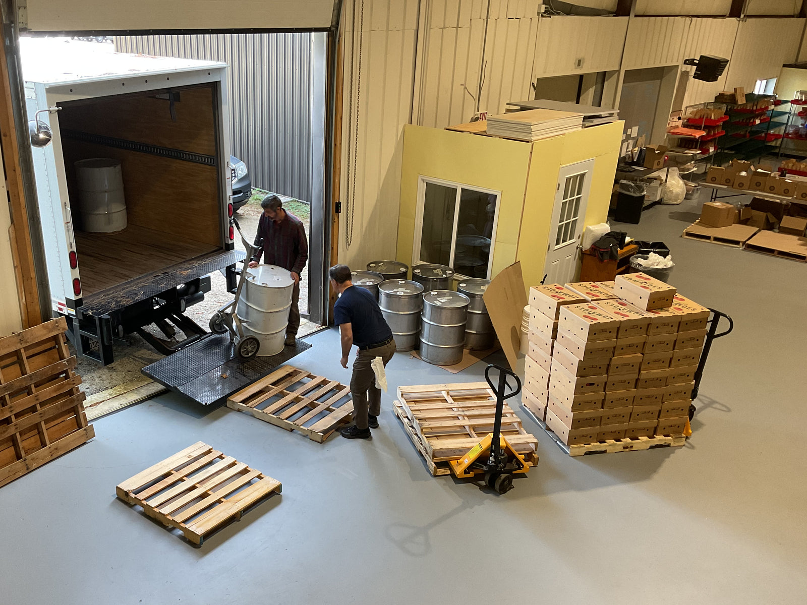 Two men load a large white barrel from a warehouse onto a truck using a metal ramp. Wooden pallets, stacked boxes, and kegs are nearby inside the spacious warehouse with a small yellow office.