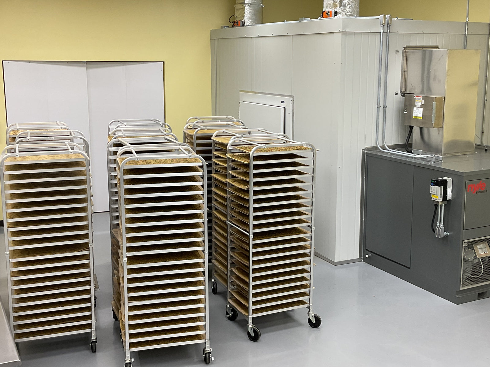 Several metal racks with trays of food are arranged in a clean, industrial kitchen or food processing facility with a large metal machine and white walls in the background.