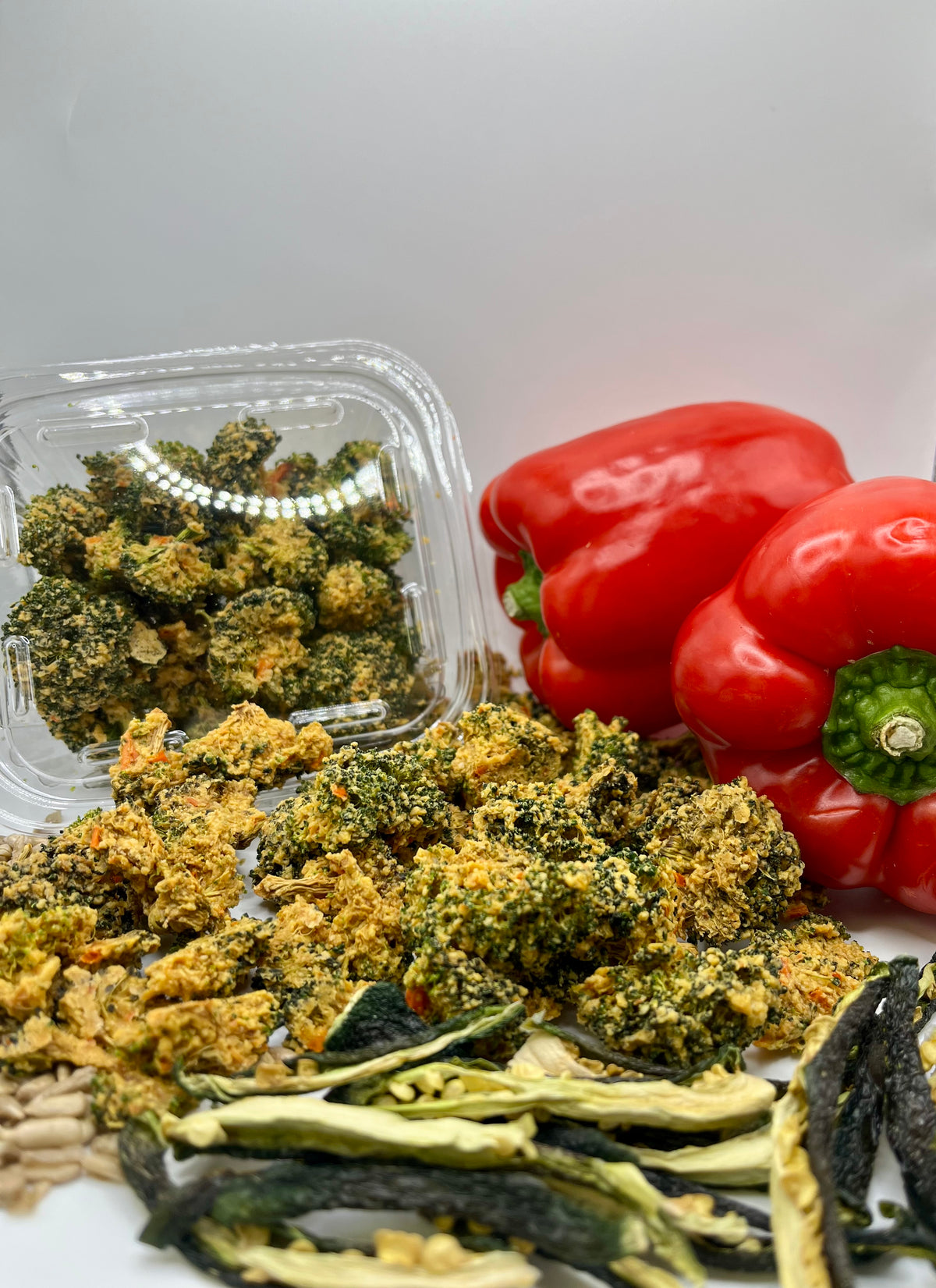 Healing Home Foods Peppy Broccoli Bites—a clear plastic container and a pile of these dried, crumbly veggie snacks—sit on a white background with fresh red bell peppers, sunflower seeds, dried zucchini strips, and a sprinkle of nutritional yeast.