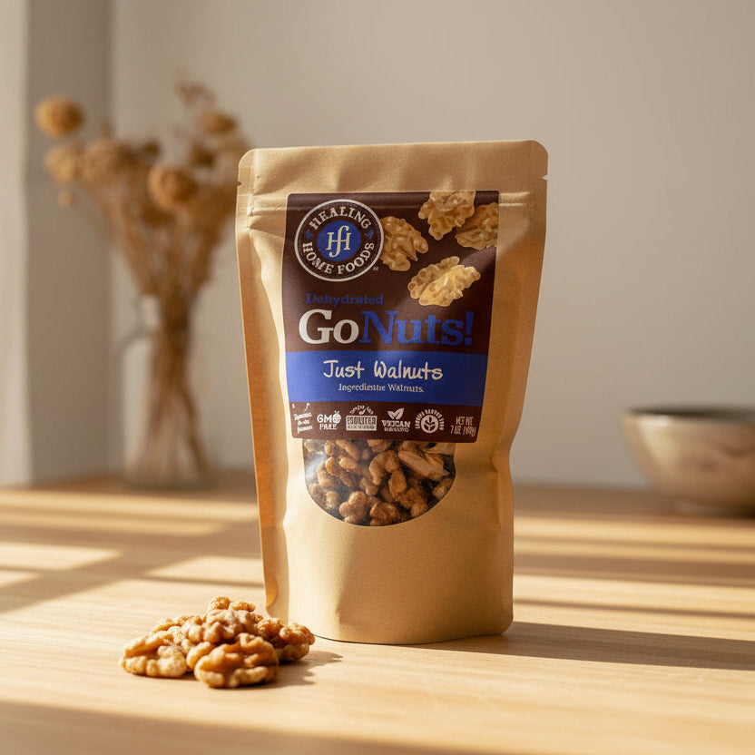 A pouch of Healing Home Foods Just Walnuts rests on a sunlit wooden table, walnut pieces scattered in front, with a blurred dried flower arrangement in the background.