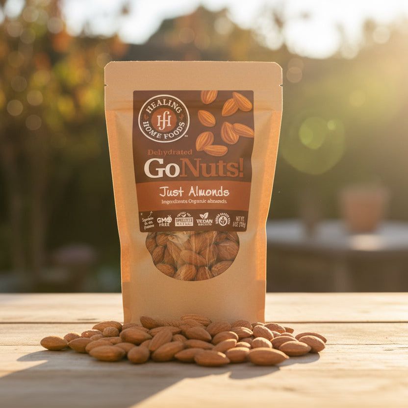 A package of Healing Home Foods Organic Just Almonds rests on a wooden table outdoors, surrounded by scattered almonds, with sunlight and blurred greenery in the background.