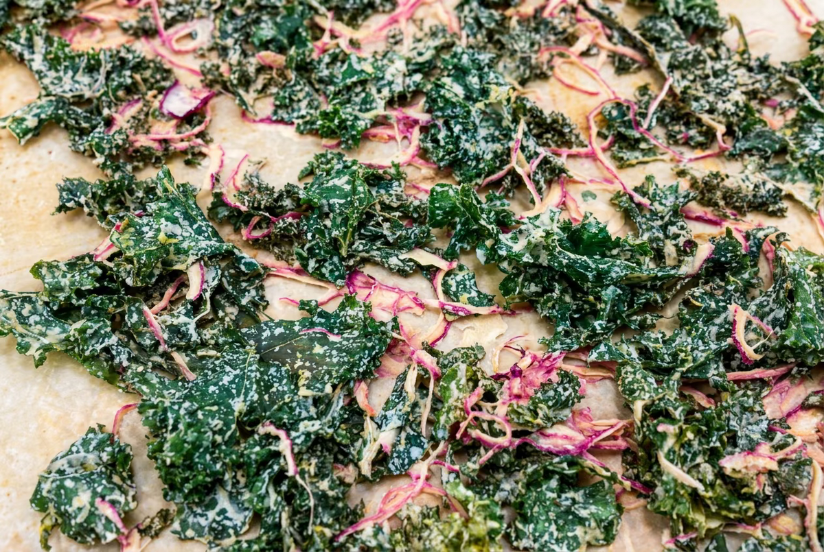 Crispy baked kale chips with pinkish-red beet stems and white cheese-like seasoning, spread out on parchment paper.