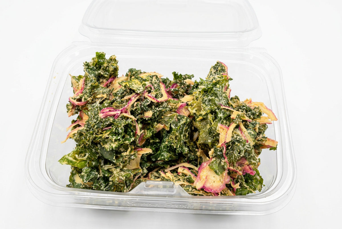 A clear plastic clamshell container filled with kale chips tossed in a creamy dressing, placed on a stainless steel countertop.
